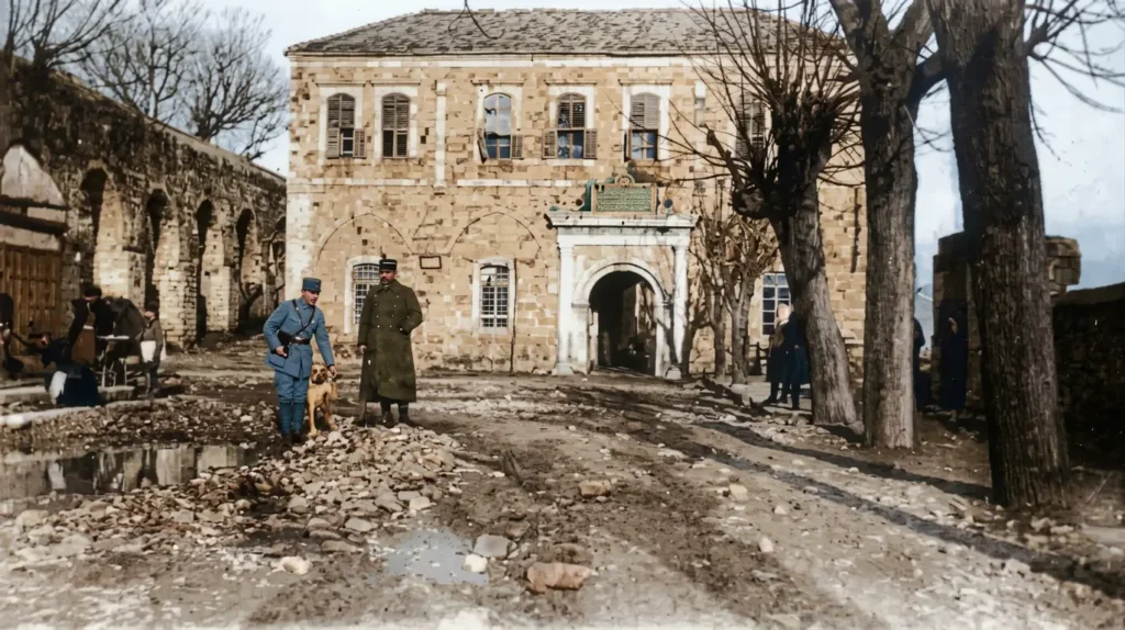 صورة تاريخية ملونة تُظهر واجهة قصر الأرناؤوط الحجري في مدينة حماة السورية، ويقف أمامه ضابطان بزي عسكري فرنسي مع كلب، والطريق أمام القصر ترابي وحجري يعود لبدايات القرن العشرين.